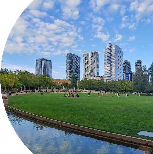 Scenic view of downtown Bellevue, featuring modern skyscrapers against a blue sky with scattered clouds, a large green park in the foreground with people enjoying the open space, and a calm water feature reflecting the sky.
