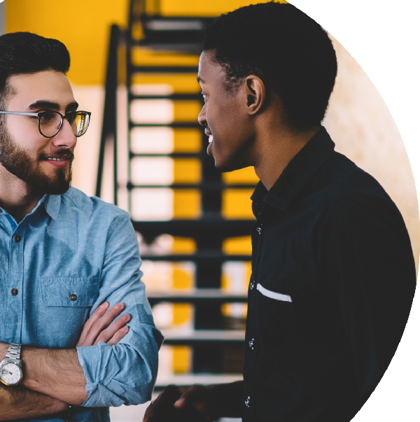 Two people are standing and having a conversation in front of a staircase. One person is smiling with arms crossed, while the other gestures as they speak.