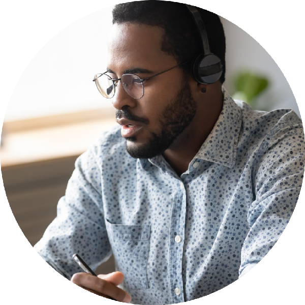 A man wearing a headset and glasses is focused while working on a computer, holding a pen, likely engaged in a virtual meeting or discussion.
